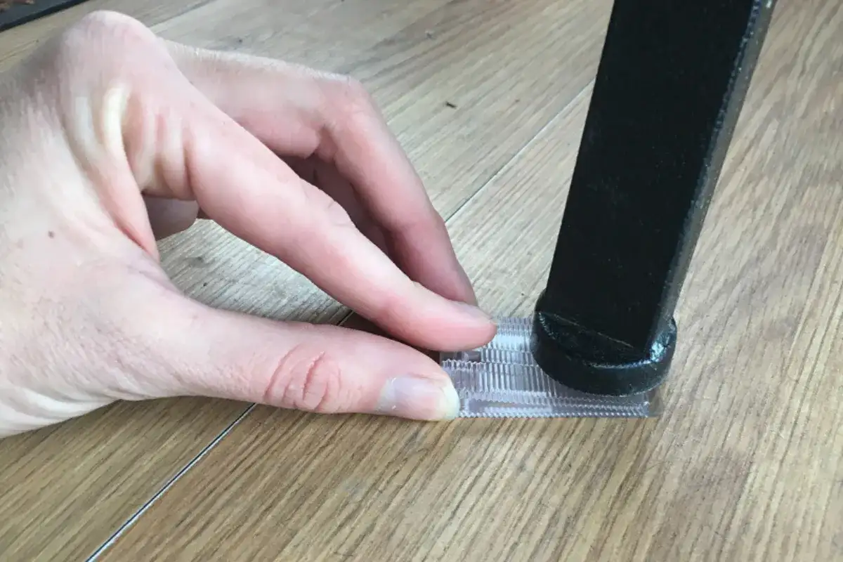 Hand placing a transparent support pad under a metal table leg on a wooden floor to level an uneven table