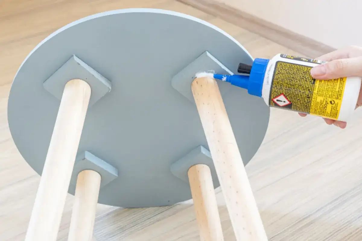Hand applying wood glue to a loose wooden table leg joint under a round tabletop