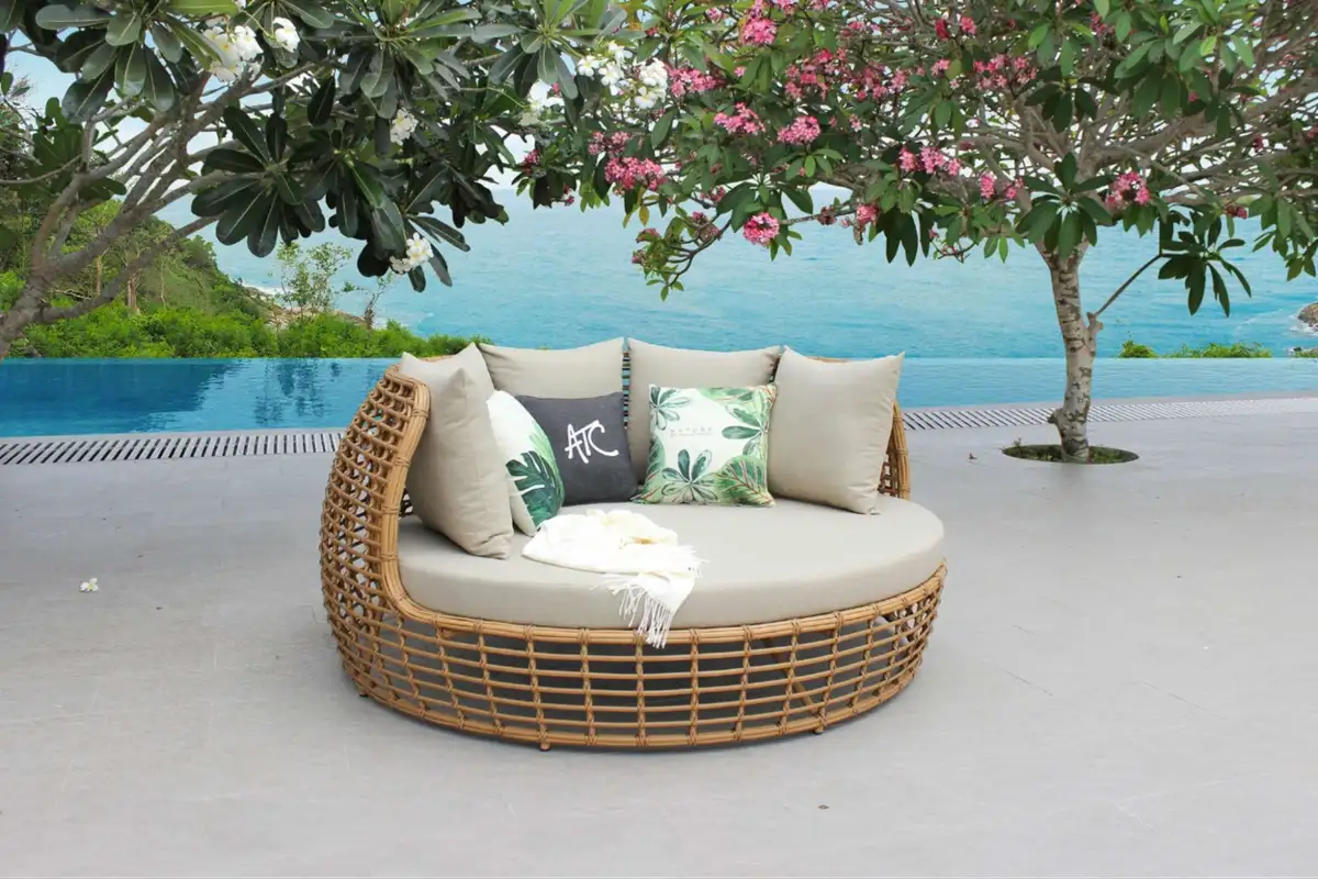 Mẫu ghế nằm tròn bằng mây, phối đệm hoa nhiệt đới, được đặt cạnh hồ bơi dưới bóng cây nở hoa.