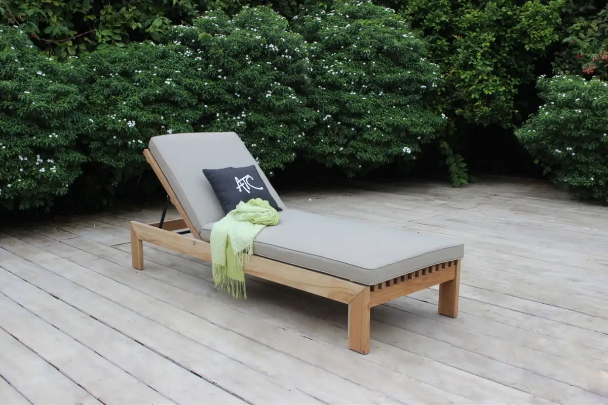 Ghế tắm nắng bằng gỗ teak với đệm màu xám và gối ATC, đặt trên sàn gỗ ngoài trời, bao quanh là hàng rào cây xanh tươi tốt.