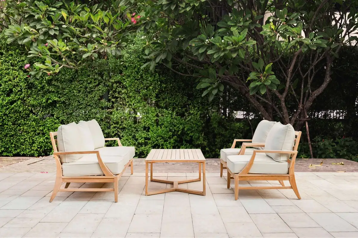 Bộ sofa ngoài trời bằng gỗ với đệm màu be, sắp xếp quanh bàn cà phê trên nền sân lát gạch, bao quanh bởi cây xanh và tán lá rợp mát.