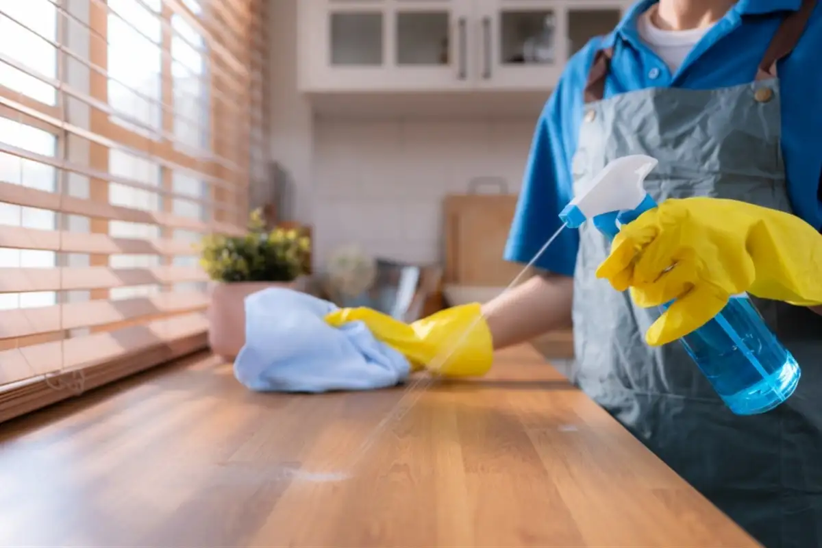 Một người đang lau chùi bề mặt gỗ bằng khăn và dung dịch xịt, đeo găng tay bảo hộ, bên khung cửa sổ có rèm sáng.
