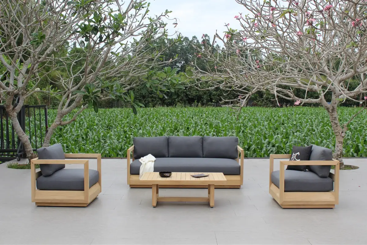 Bộ ghế sofa gỗ ngoài trời kèm đệm tối màu đặt trên nền gạch, bao quanh là cây trụi lá và cánh đồng xanh phía sau.