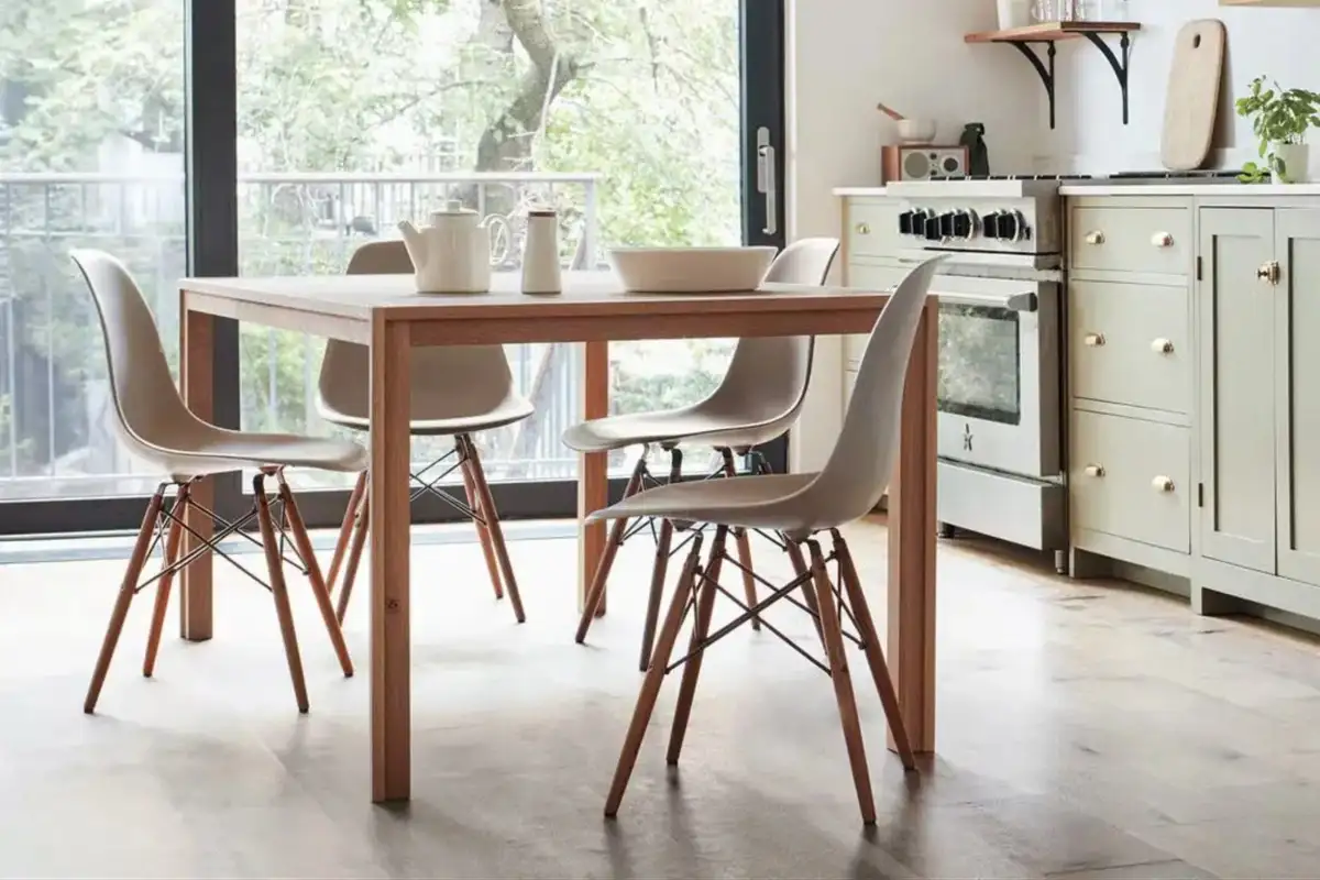 A minimalist dining space featuring a wooden dining table, white plastic chairs with wooden legs, and a light-filled kitchen backdrop.