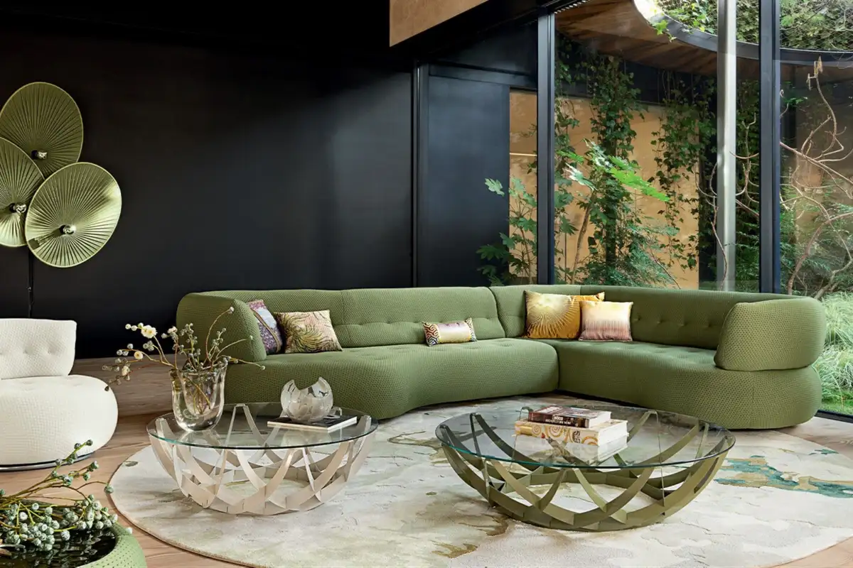A bold, eclectic interior featuring a curved green sofa, artistic glass coffee tables, playful decor, and large windows framing a lush garden view.
