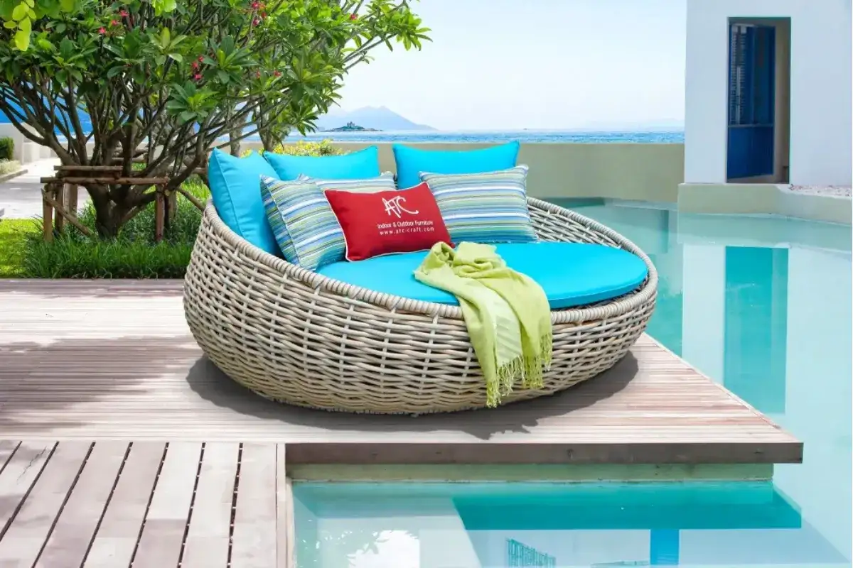 Giường tắm nắng mây nhựa hình tròn LAIKA với đệm xanh biển và gối trang trí đặt trên sàn gỗ cạnh hồ bơi phong cách resort