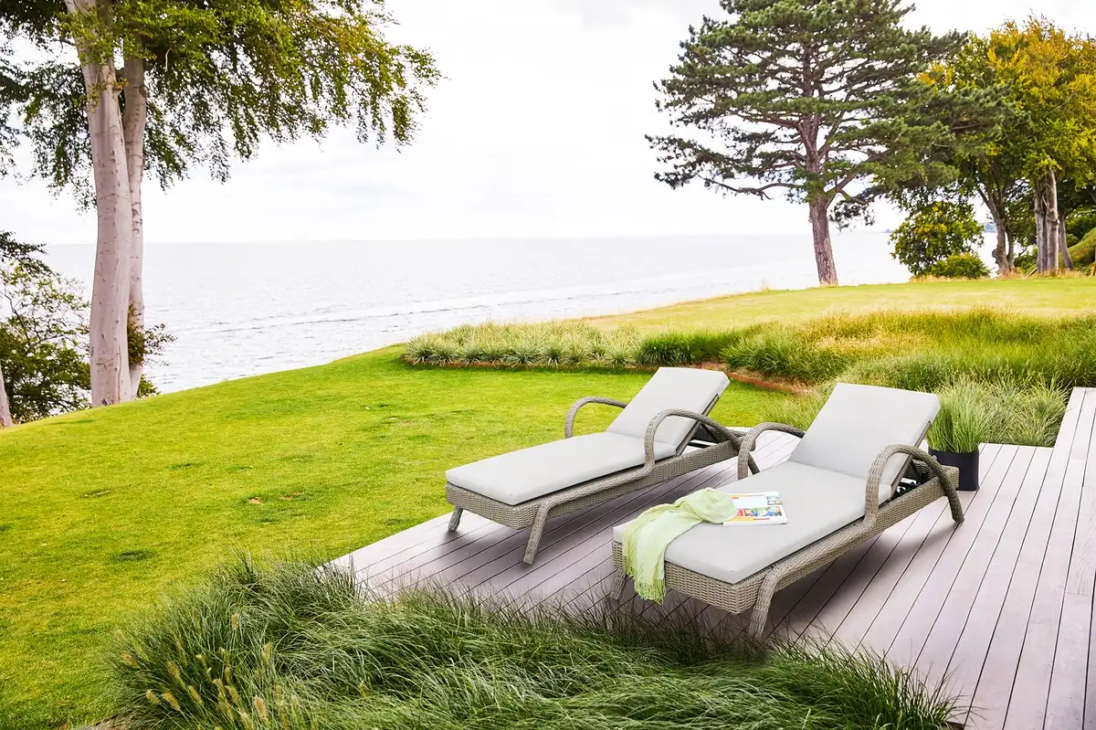 Ghế tắm nắng ngoài trời đặt trên thảm cỏ ven biển, thiết kế mây nhựa đan kết hợp đệm xám nhạt tạo không gian thư giãn hòa hợp thiên nhiên.  