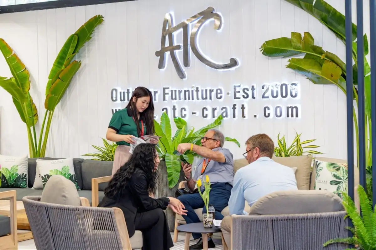 Khách hàng quốc tế trao đổi cùng nhân viên tại gian hàng ATC Furniture trong triển lãm, không gian trưng bày hiện đại với logo thương hiệu nổi bật và cây xanh trang trí. 