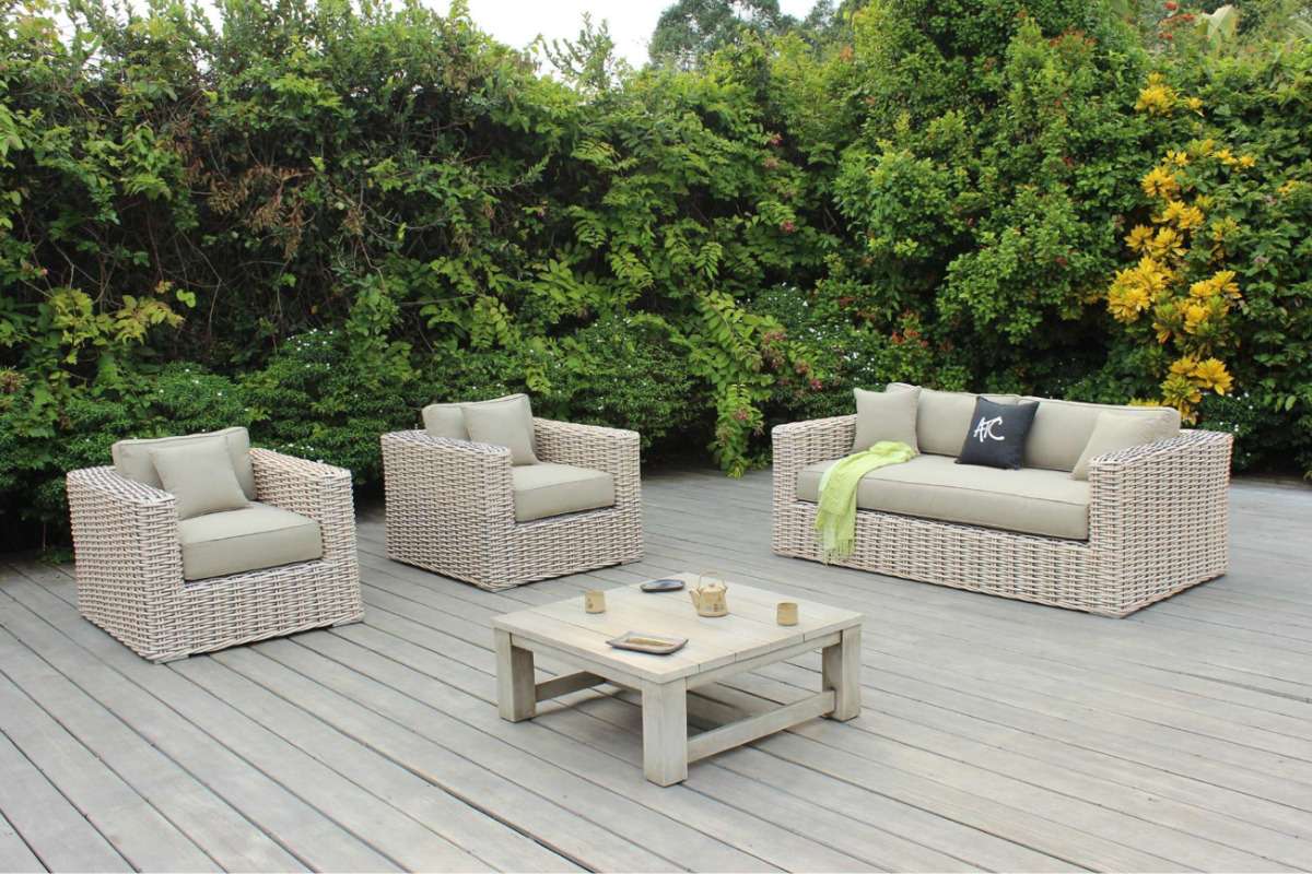 Neutral-tone rattan outdoor sofa set with clean lines and simple silhouettes arranged on a deck in front of lush greenery.