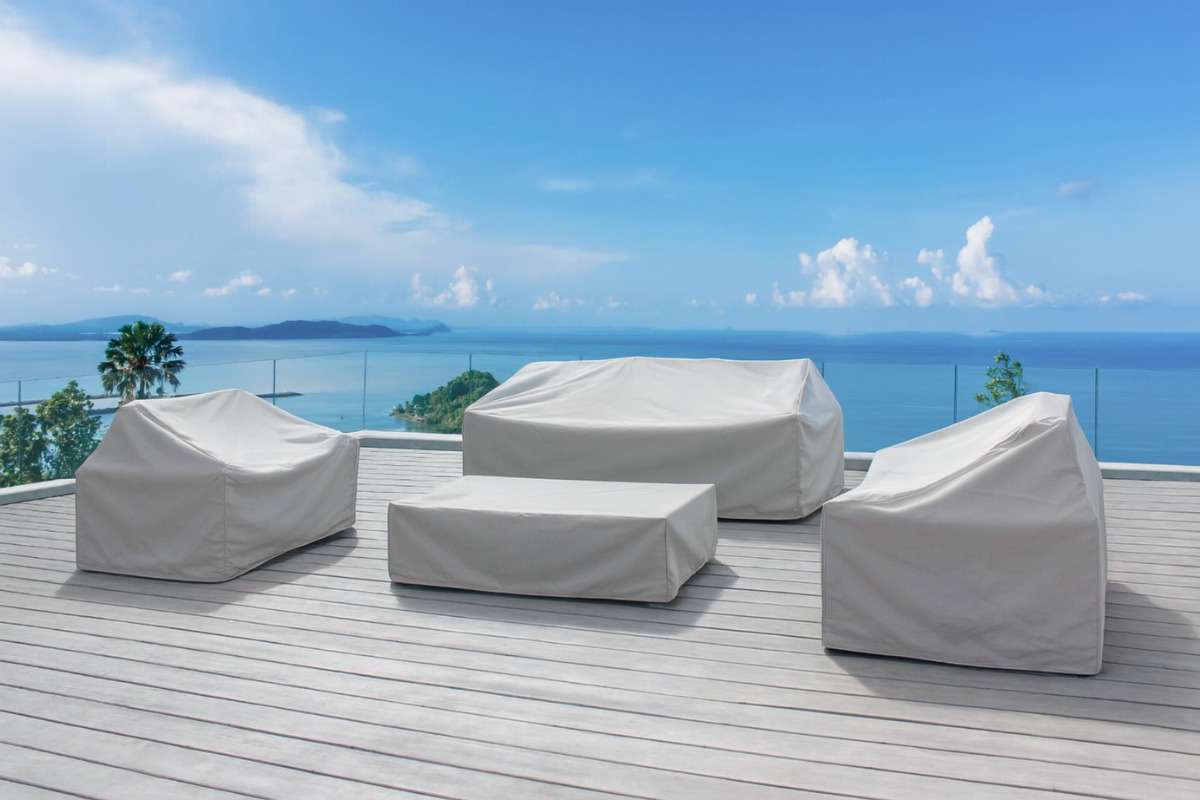 Outdoor sofa and chairs fully covered with protective covers on a coastal deck, illustrating seasonal care in rainy and high-UV environments.
