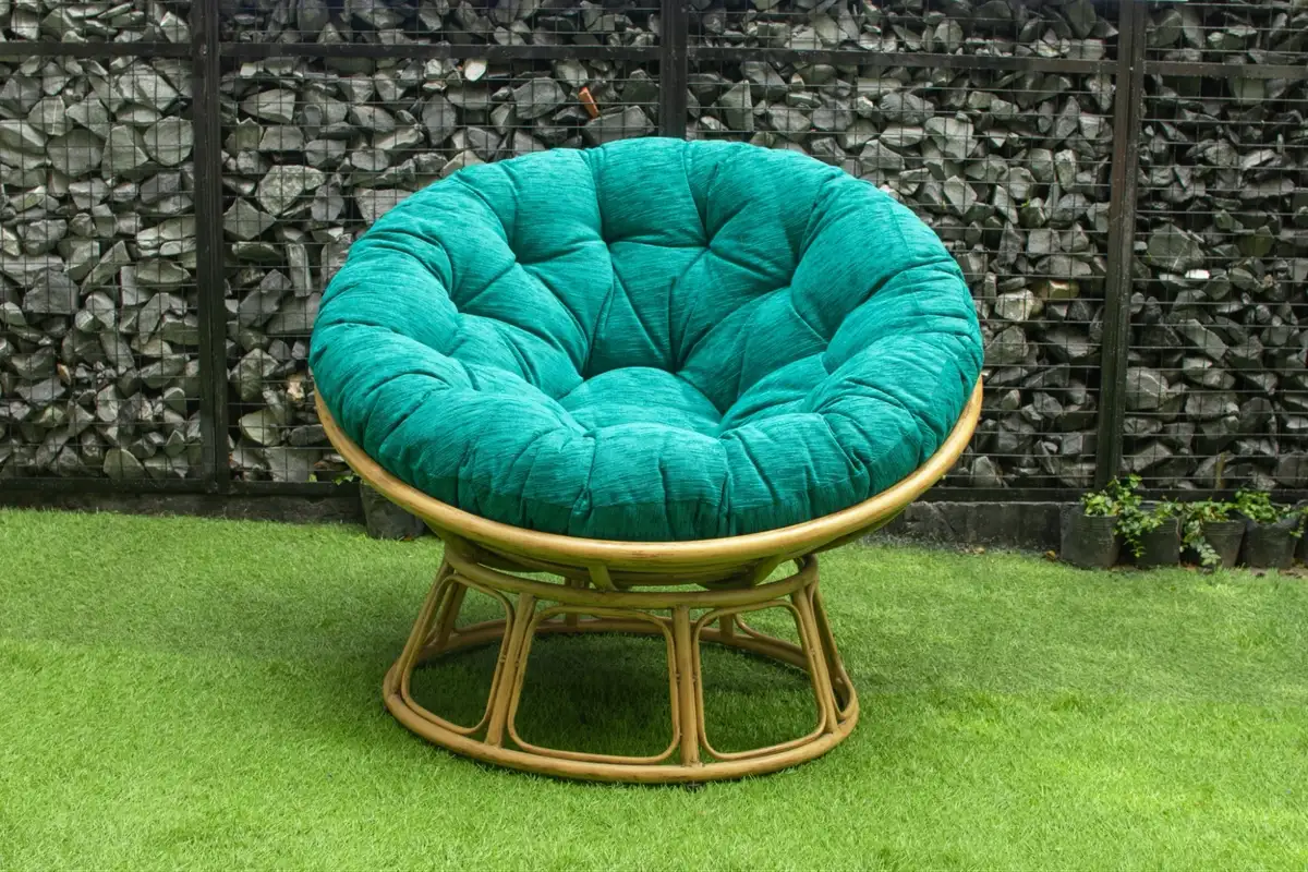 Green cushioned papasan chair with a rattan base placed on artificial grass against a stone wall.