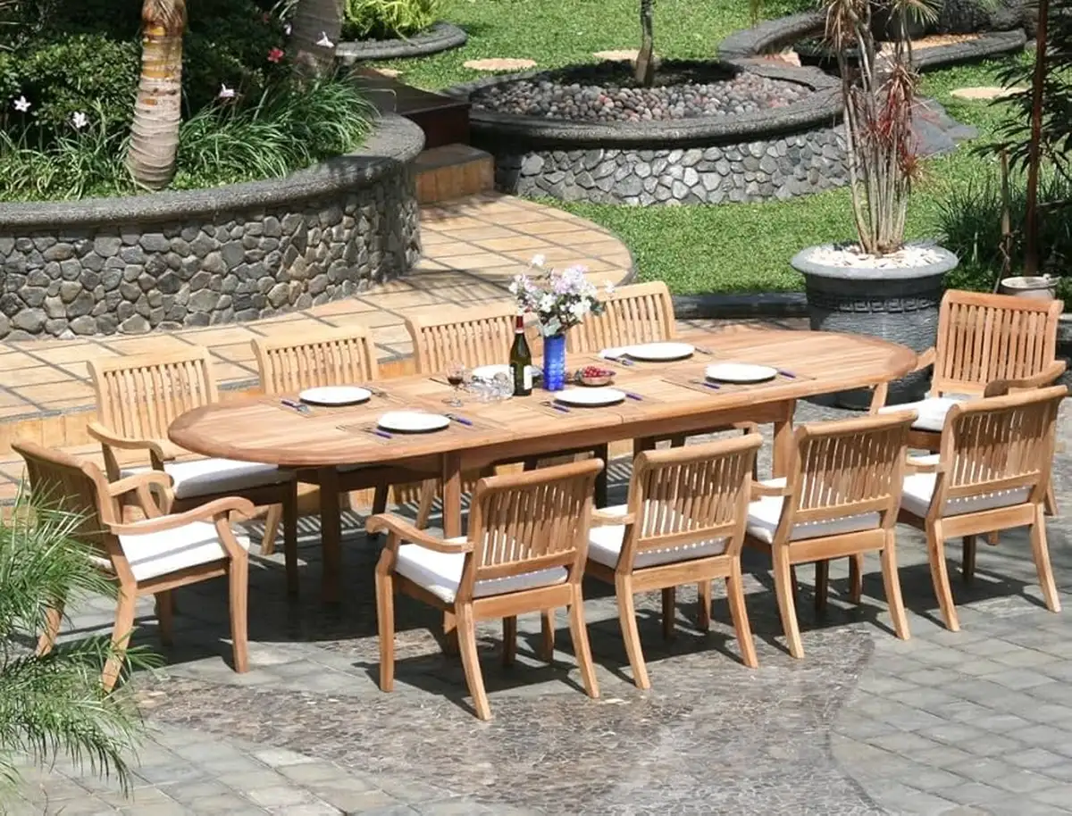 Large outdoor teak dining table set with wooden chairs and white cushions arranged in a landscaped garden