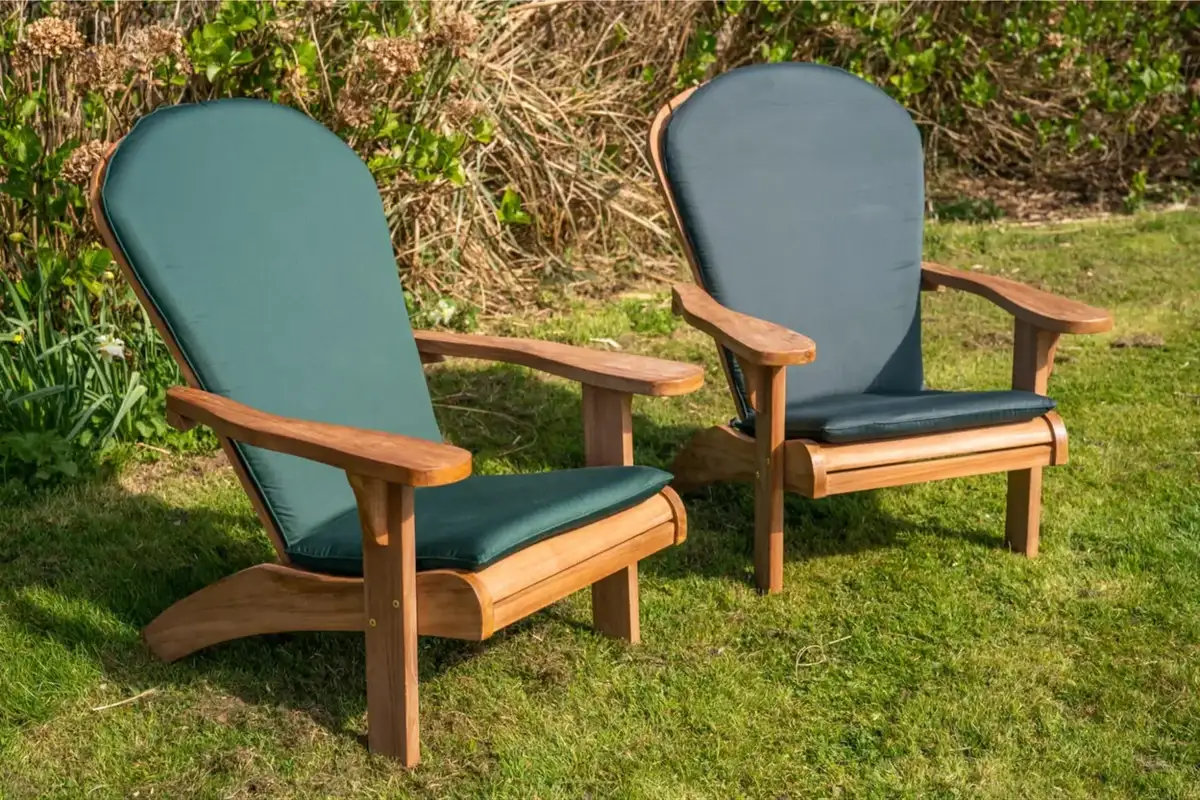 Two wooden Adirondack chairs with green weather-resistant cushions placed on grass in a garden setting.  
