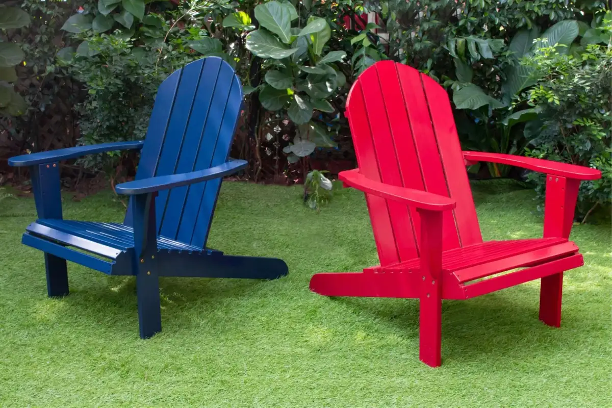 Red and blue Adirondack chairs made from recycled plastic, placed on green lawn with garden plants in the background.  