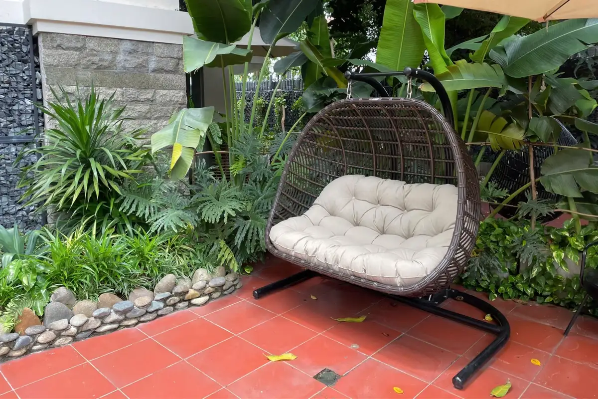 Brown rattan swing chair with plush seating placed under banana leaves and beside river stones. 