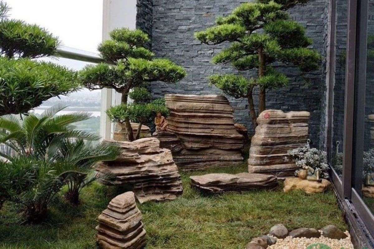 Small Zen garden with layered natural rocks, trimmed pine trees, grass, and a gray stone wall