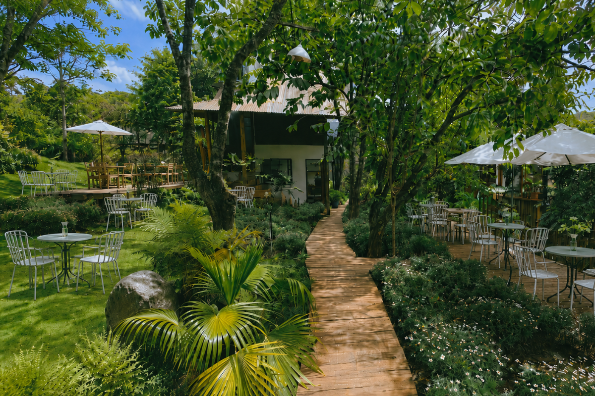 Lush garden café with wooden walkways, outdoor tables, white chairs, umbrellas, and tropical greenery