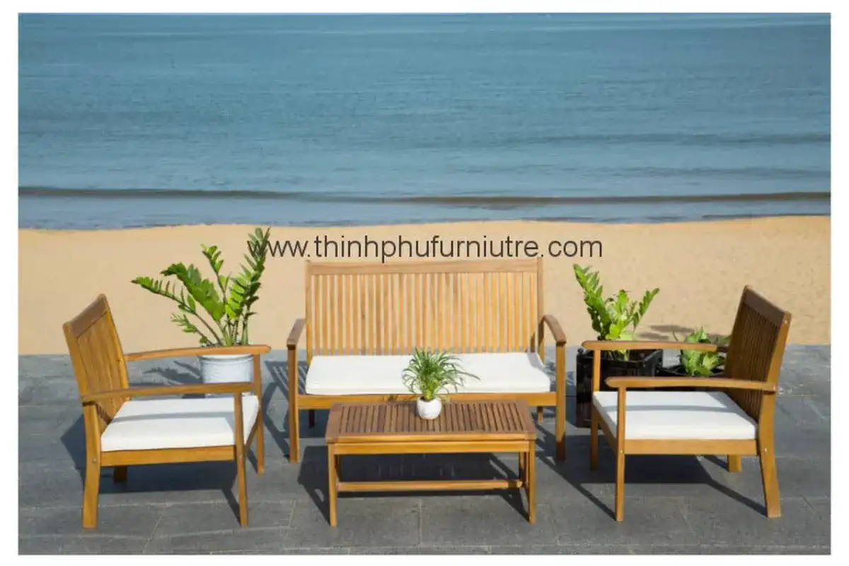 Wooden outdoor furniture set by Thinh Phu, including a bench, chairs, and a coffee table with white cushions, displayed by the beachside.
