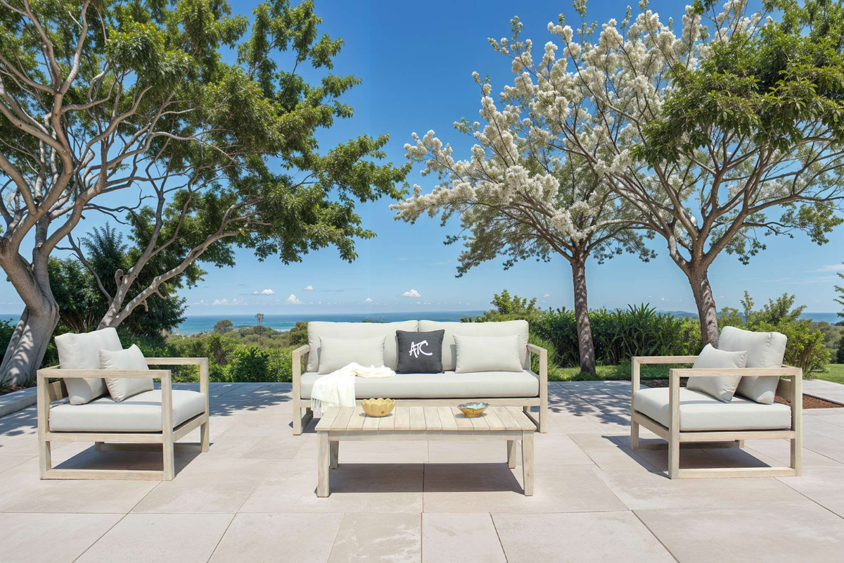Bộ sofa khung gỗ đặt ngoài trời với nệm sáng màu, bố trí trên sân lát đá dưới hàng cây và hướng nhìn ra biển.