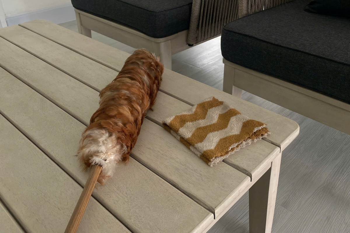 Bàn gỗ ngoài trời với cây chổi lông và khăn lau đặt trên bề mặt, minh họa dụng cụ dùng để vệ sinh định kỳ.