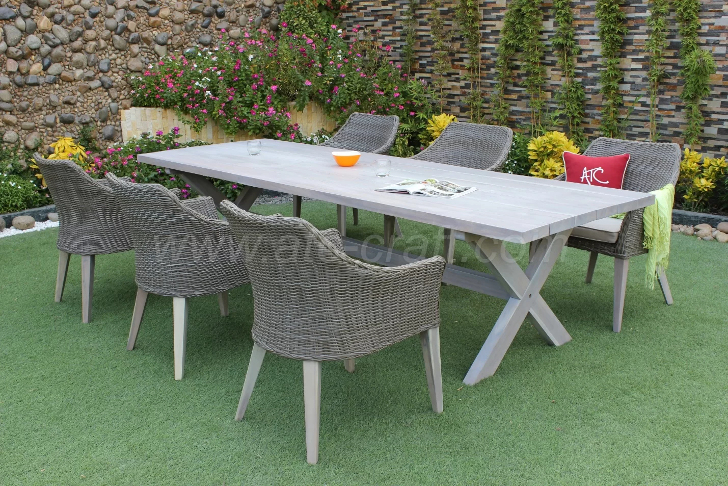 A stylish outdoor dining set featuring a long wooden table and six wicker chairs, arranged on artificial grass with a backdrop of flowers and a decorative stone wall