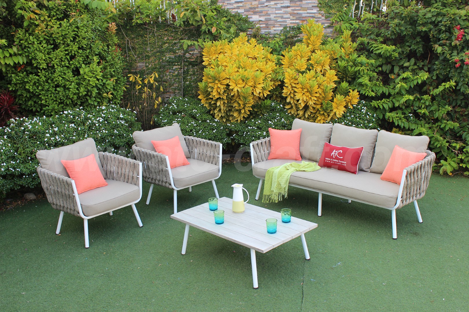 A modern outdoor wicker conversation set with a sofa, two armchairs, beige cushions, and coral accent pillows, placed in a lush garden with green shrubs and yellow plants