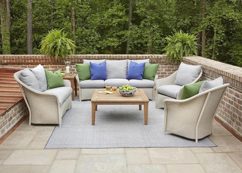 Outdoor seating area with white wicker furniture, colorful pillows, and a wooden coffee table, set against a brick wall and surrounded by greenery