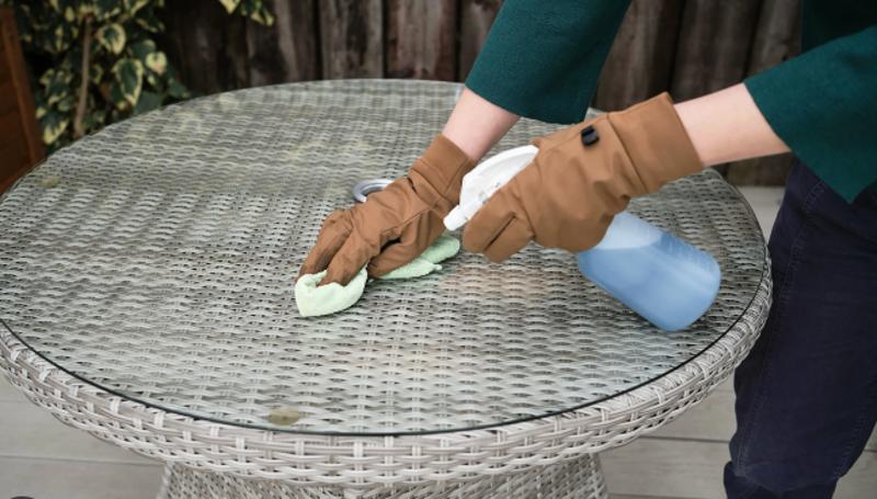 Person cleaning a wicker outdoor table with gloves, using a spray bottle and cloth to maintain the surface, emphasizing proper patio furniture care