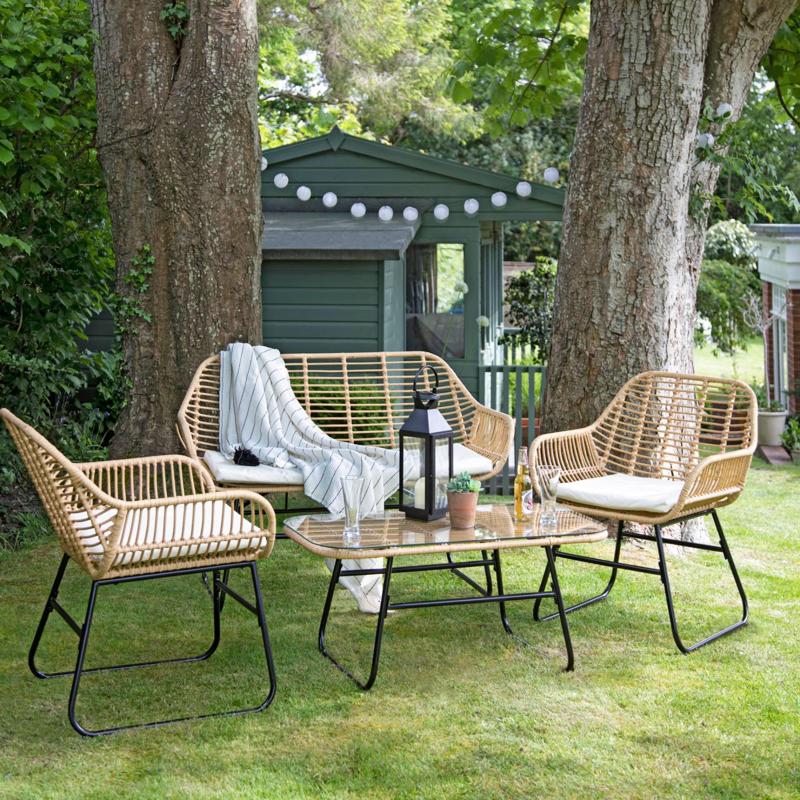 Charming outdoor seating area with a wicker sofa, chairs, and glass table, set beneath large trees, creating a cozy garden space with string lights and lantern decor