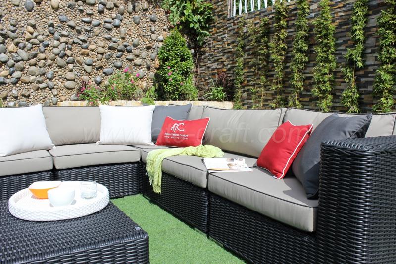 A modern outdoor rattan sectional sofa set with neutral beige cushions, red and gray accent pillows, placed on artificial grass in a garden setting with a decorative stone wall and vertical greenery