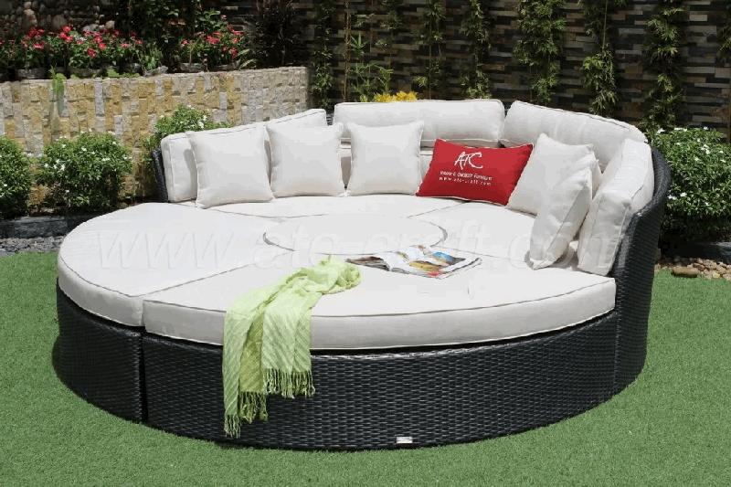 Round outdoor daybed with white cushions and a red ATC pillow, set on a grassy patio surrounded by greenery and decorative stone walls