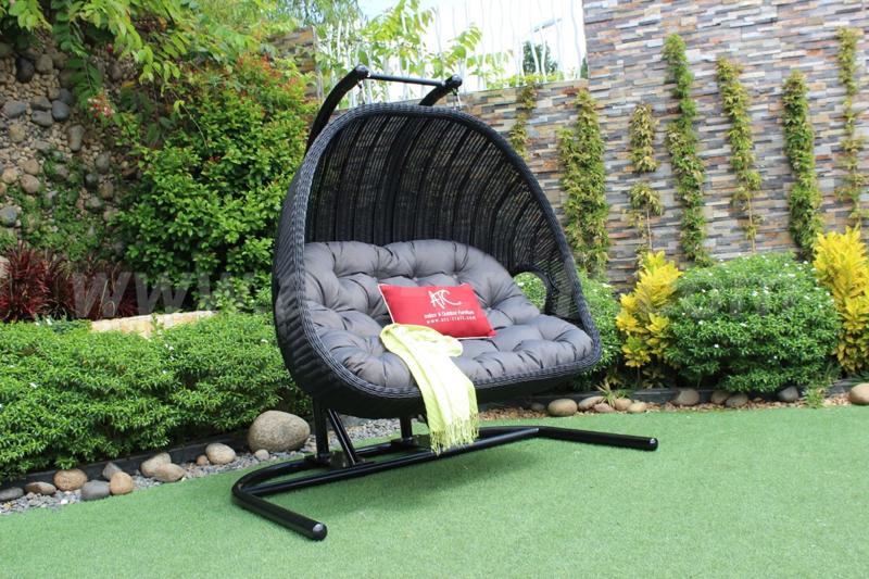 Outdoor wicker hanging chair with comfortable cushion, placed on green artificial grass in a garden setting. Decorated with a red ATC Furniture pillow and yellow throw blanket